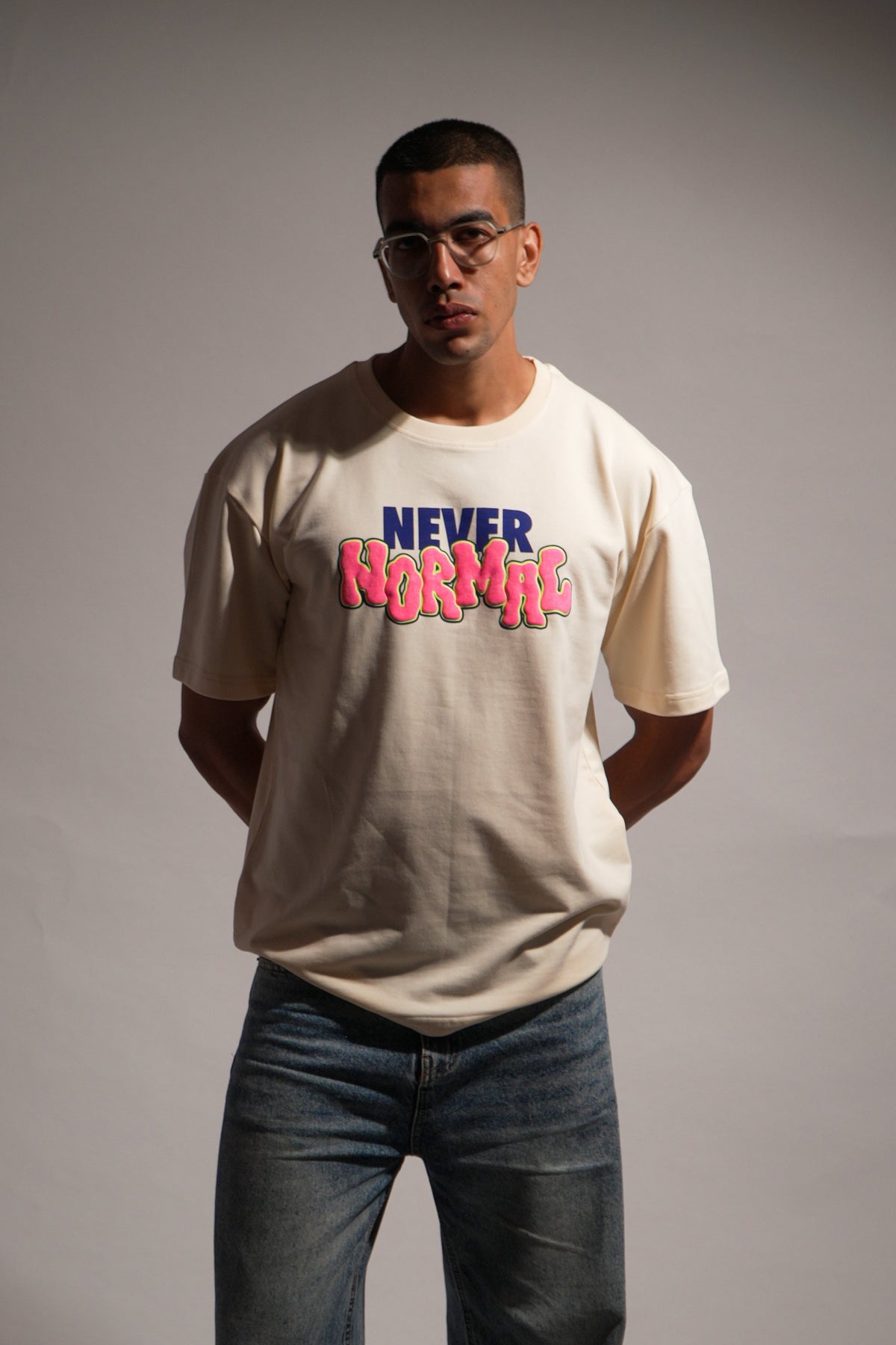 A man wearing a cream-colored t-shirt with a colorful 'NEVER BOTHER' graphic in blue and pink, paired with dark blue jeans and gray sneakers, standing against a gray studio background.